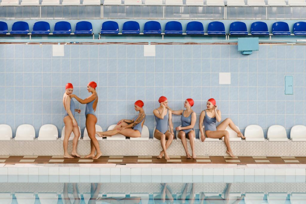 Competitive swimmers wearing matching one-piece swimsuits and swim caps sitting and standing along an indoor pool, representing swimwear production supported by a swimwear ERP system.